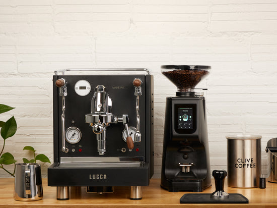 Matte black LUCCA M58 espresso machine with the LUCCA Atom 75 espresso grinder in black with LUCCA accessories: milk pitcher, tamping mat, tamper, and Clive Coffee airscape canister