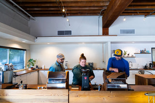Three coworkers brew coffee at a wood counter with espresso machines under exposed ceiling beams