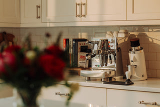 Shiny espresso machine on a kitchen counter beneath white cabinets with mugs and books nearby.