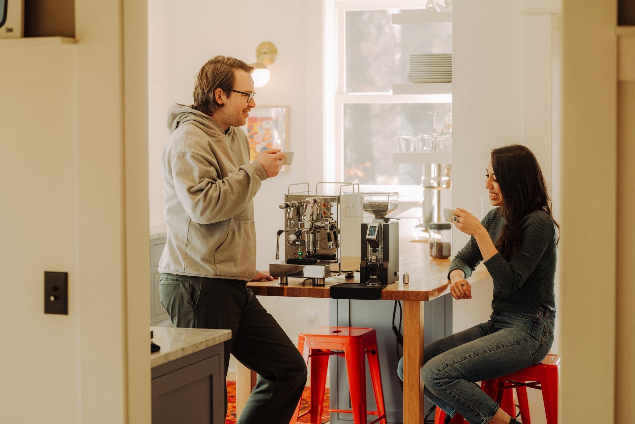 Man in glasses and hoodie stands at a cafe counter beside a two-group espresso machine while a woman sits nearby