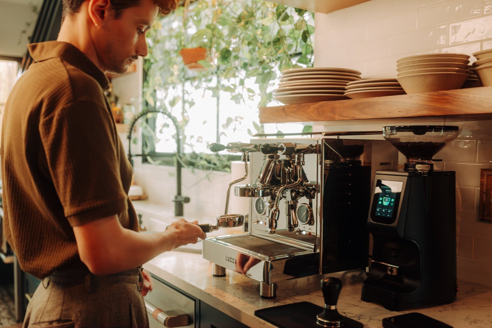 Barista in a brown sweater loads a portafilter on a shiny espresso machine with stacked plates and green plants in the background.