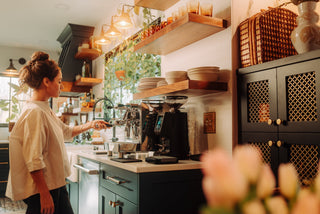 Barista in a light shirt operates a shiny espresso machine behind a cafe counter with wooden shelves and greenery.