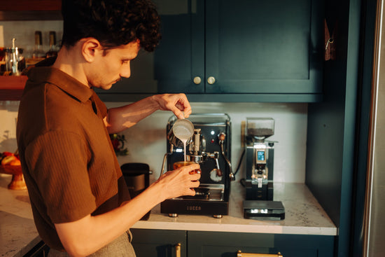 Barista in a brown shirt prepares coffee at a workstation with an espresso machine and grinder in the background.