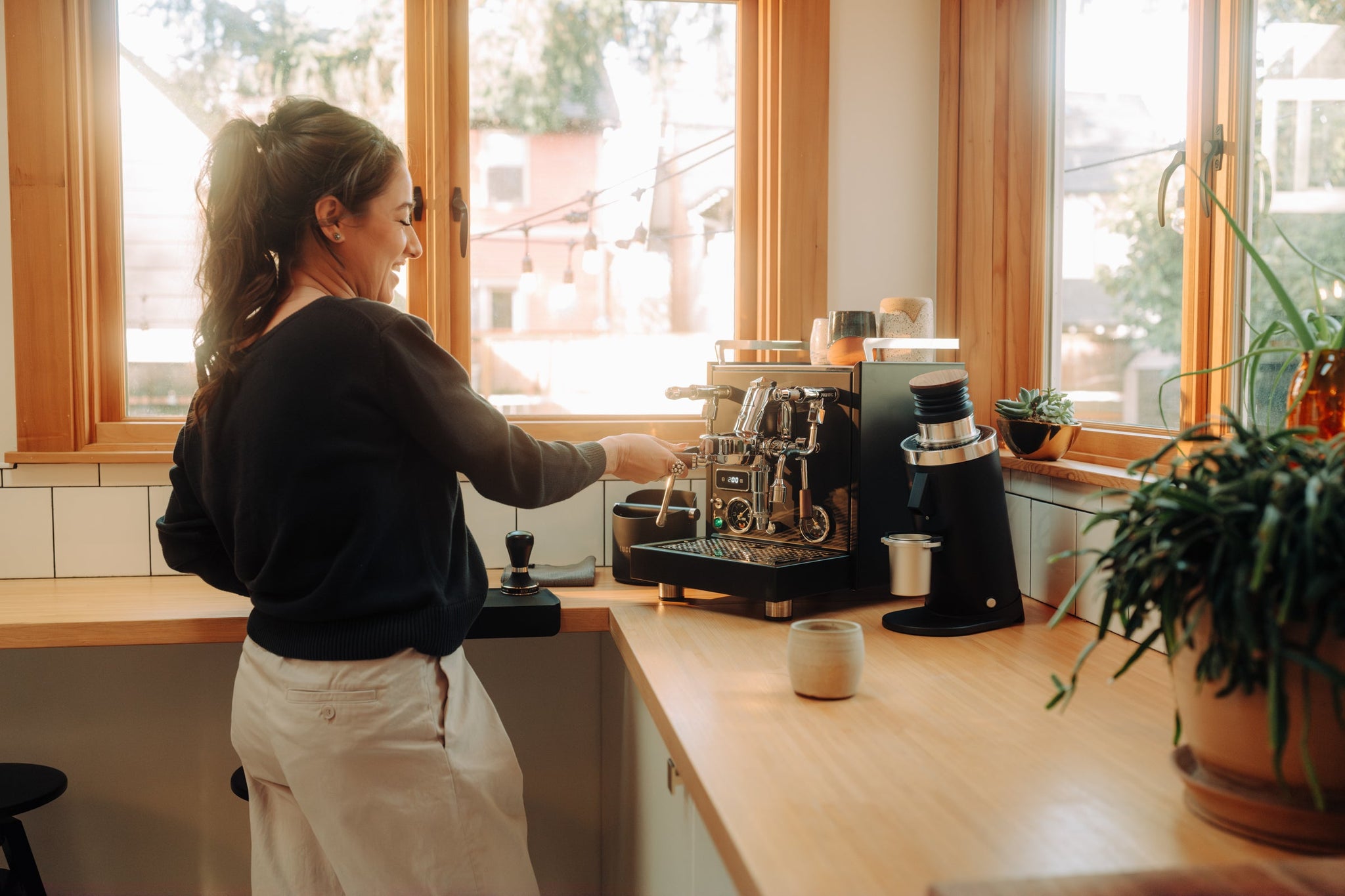 Barista in a sunlit kitchen tending an espresso setup with a grinder by the window