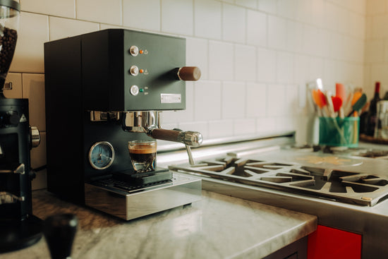Profitec GO espresso machine with wooden portafilter and a glass of espresso on the drip tray against a white tiled backsplash.