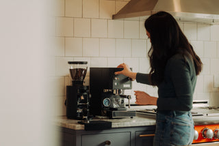 A person operating a black home espresso machine with a grinder on a white tile kitchen counter