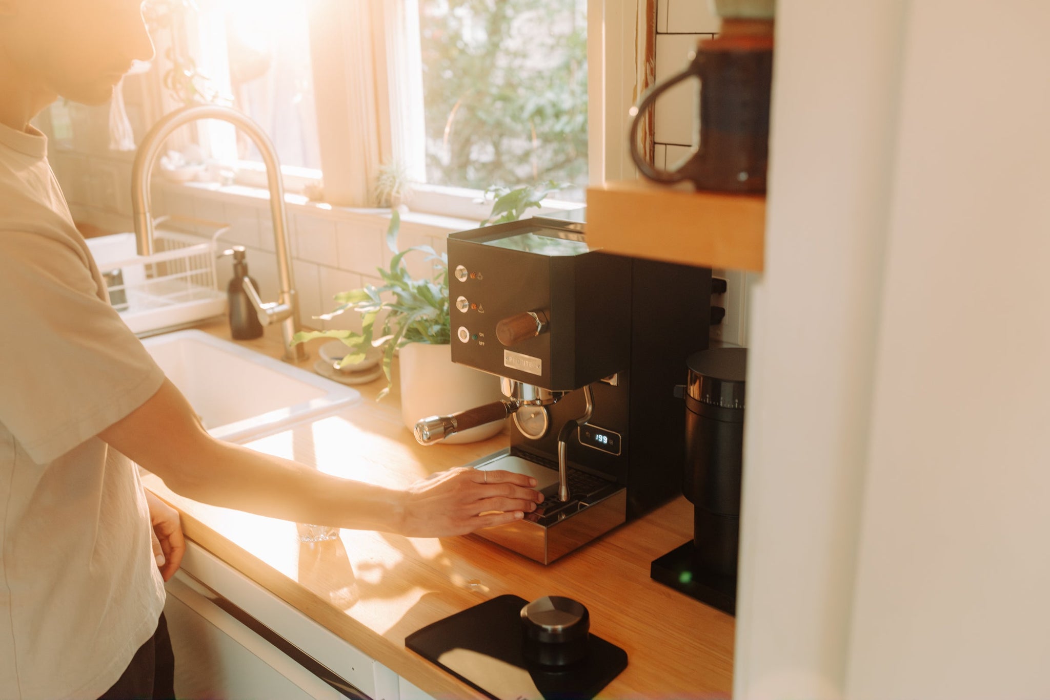 Person using a compact home espresso machine in a sunlit kitchen with a sink and plants nearby.