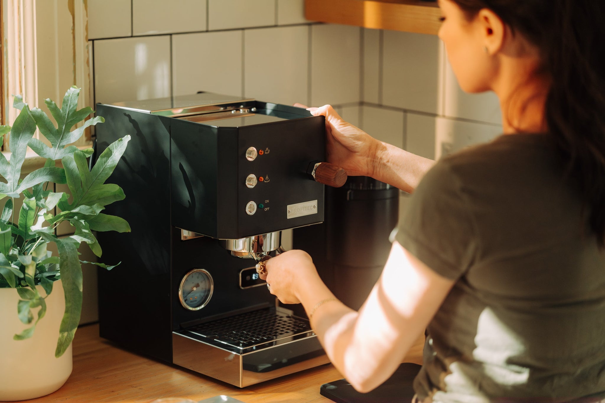 A person uses a black home espresso machine on a wood counter with a green potted plant nearby.