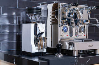 Eureka Mignon Zero grinder with clear hopper beside a shiny LUCCA espresso machine on a dark countertop