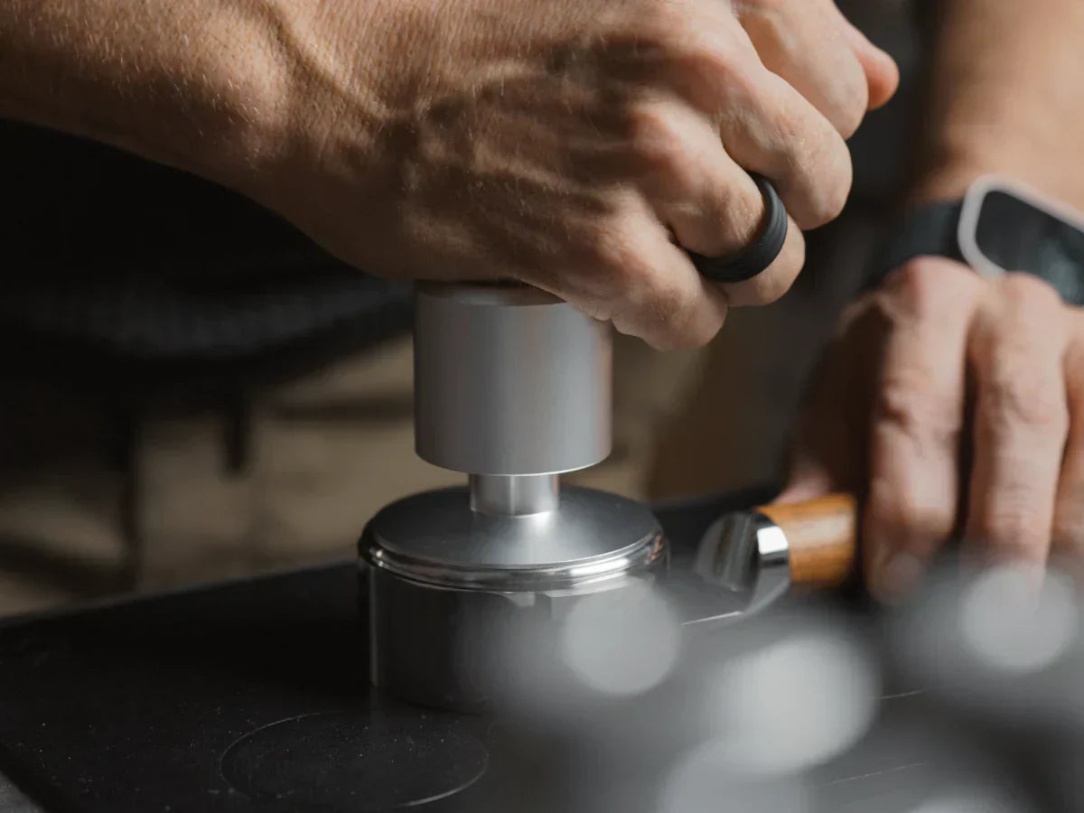 Hands tamping espresso grounds in a portafilter with a metal tamper on a dark countertop