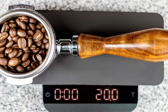 Roasted coffee beans fill a metal grinder next to a wooden handled portafilter and a digital scale