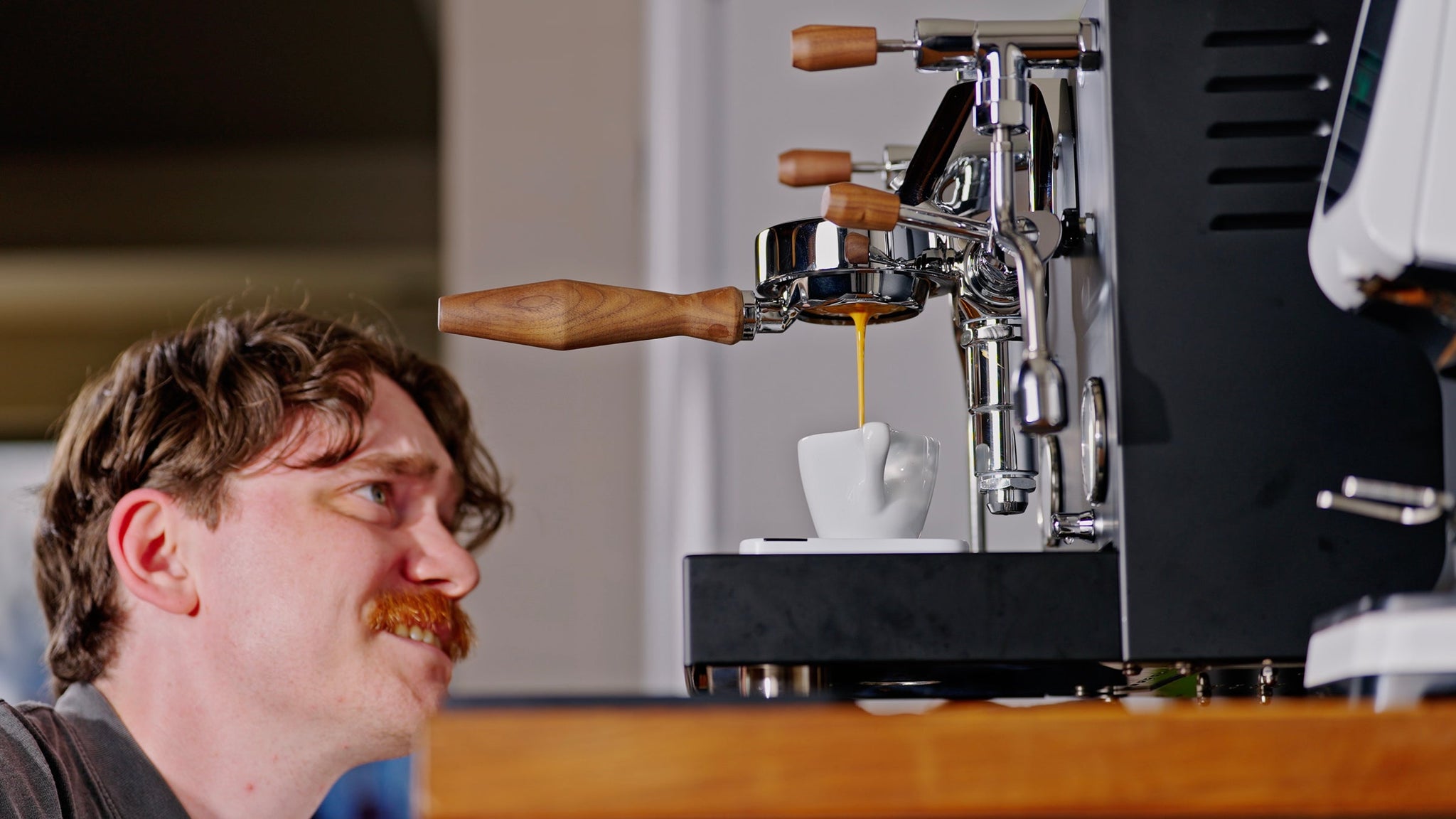 Bearded barista watches espresso drip from a portafilter into a white cup beside a high end espresso machine