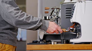 Hands operate a commercial espresso machine with wooden portafilter handles.