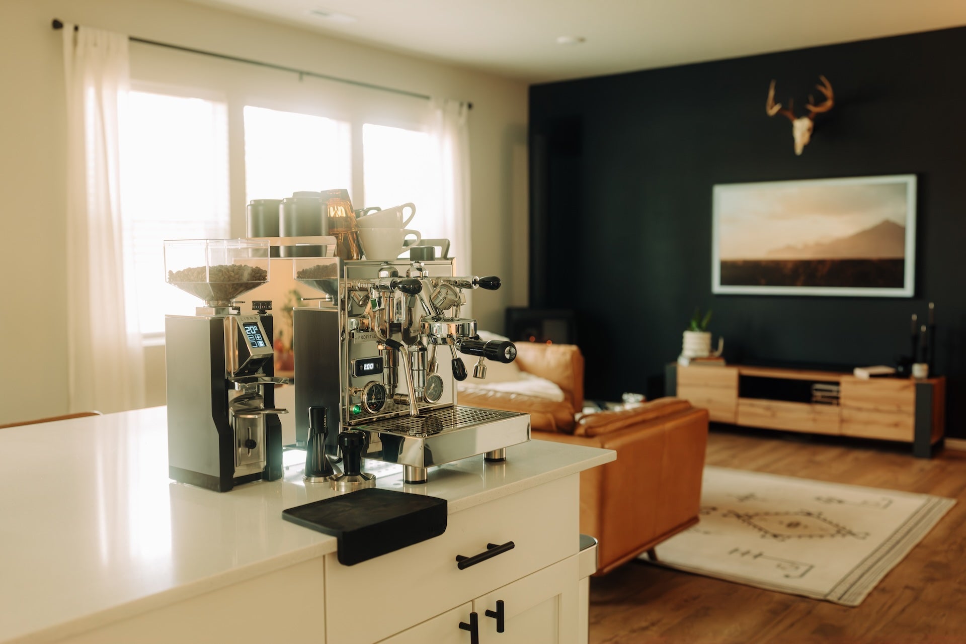 Stainless espresso machine on a white kitchen island with cups and grinders in a sunlit modern living area.