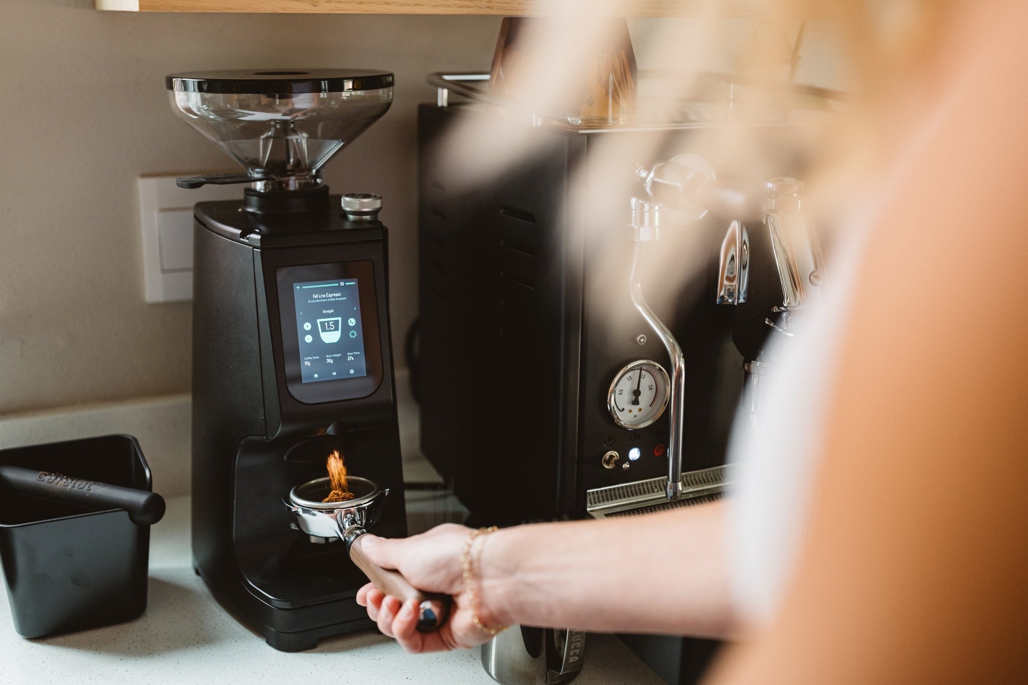 Hands hold a portafilter under a grinder next to a polished espresso machine