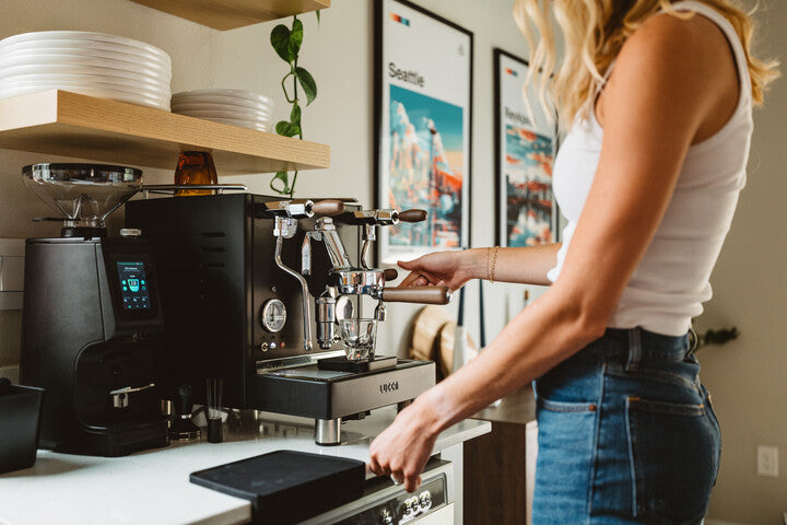 Woman hands a cup of coffee to a child at a home espresso setup