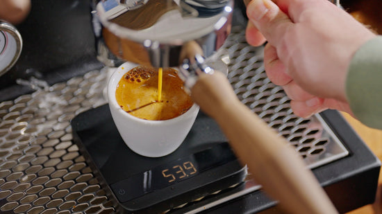 Barista pours espresso into a white cup placed on a digital scale for precise weighing.