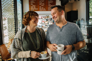 Smiling woman and man in the Cartel Roasting Co cafe, each holding a coffee cup.