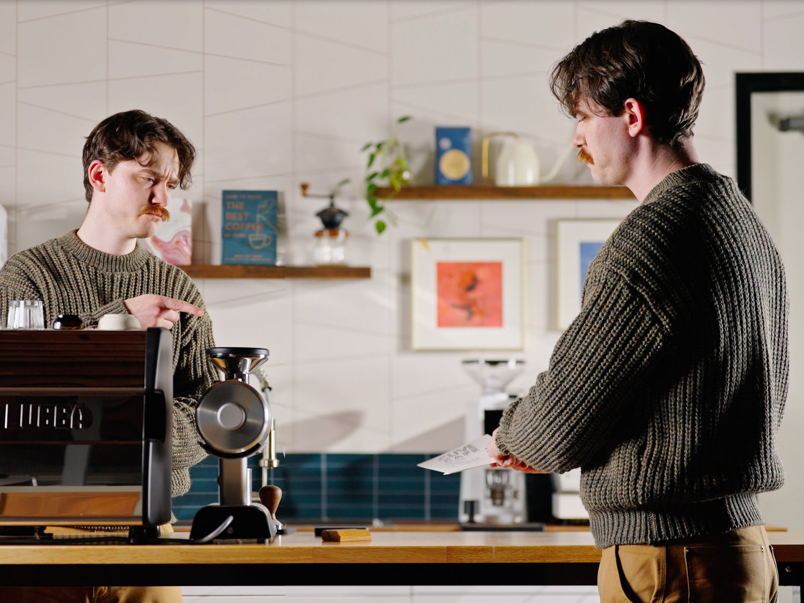 Two men with mustaches in knitted sweaters stand at a cafe counter conversing beside a coffee setup.