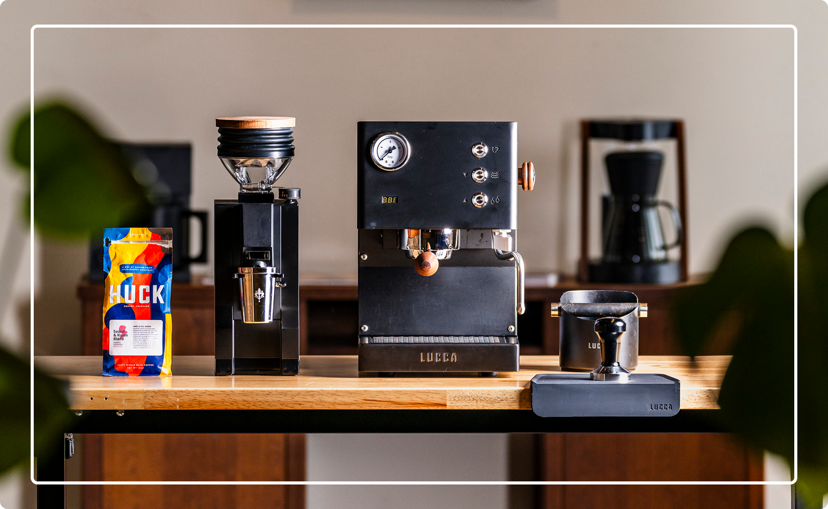 Black espresso machine with gauge beside a tall grinder, a colorful HUCK coffee bag, and a small scale on a wooden counter.