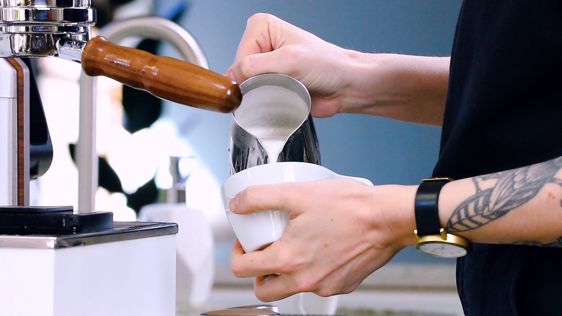 Barista with a tattooed forearm pours steamed milk from a metal pitcher beside an espresso machine.