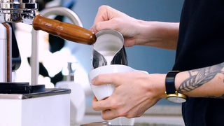 Barista frothing milk with a steam wand into a stainless steel pitcher.
