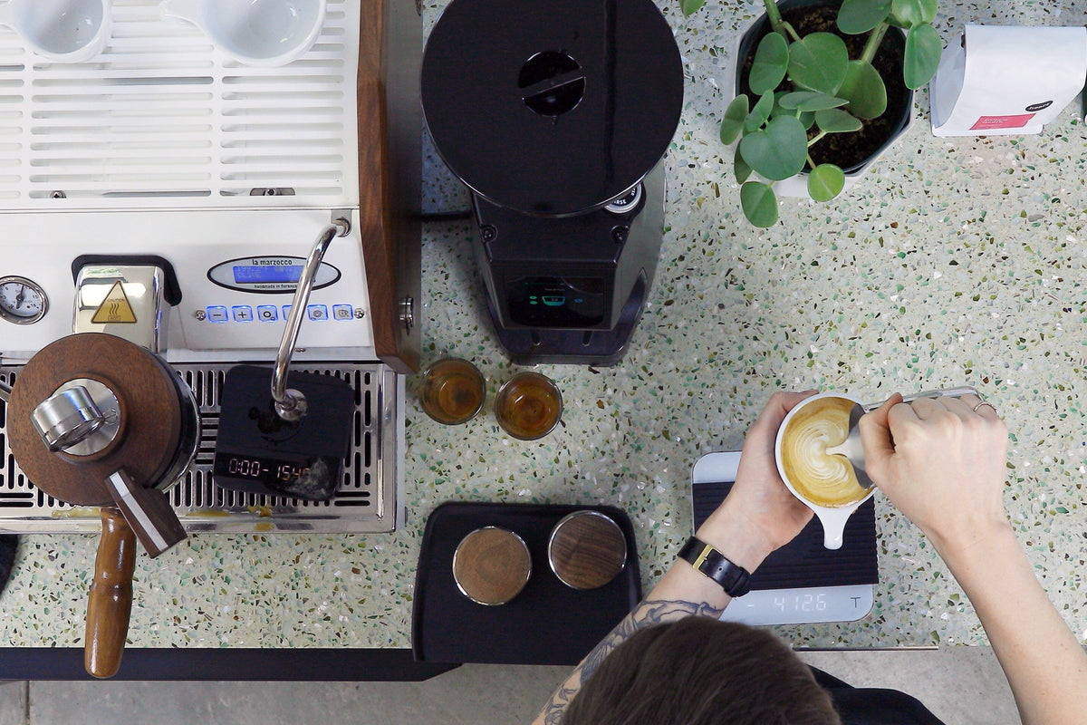 Overhead view of a coffee bar with a white espresso machine on the left, a black grinder center, a potted plant on the right, and a person making a latte.