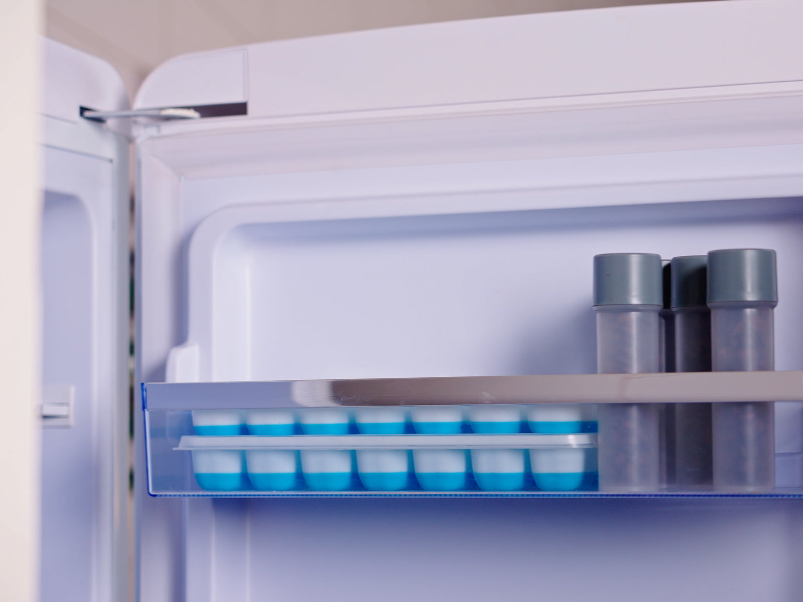 Open refrigerator interior with shelf holding blue ice cube trays and gray bottles
