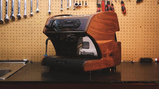 Espresso machine with a custom wooden shell sits on a workshop workbench with pegboard and tools in the background.