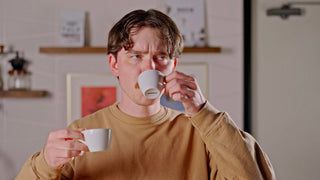 Man in a tan sweatshirt sips from a white espresso cup while holding another cup