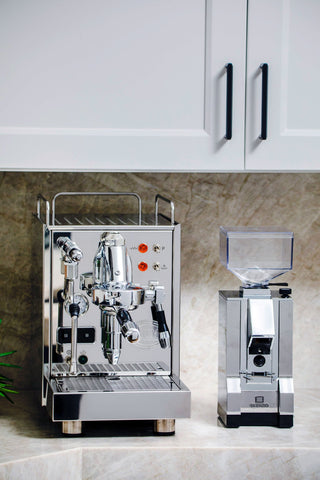 Shiny chrome espresso machine with dual portafilters on a marble countertop beside a tall grinder, against white kitchen cabinets.