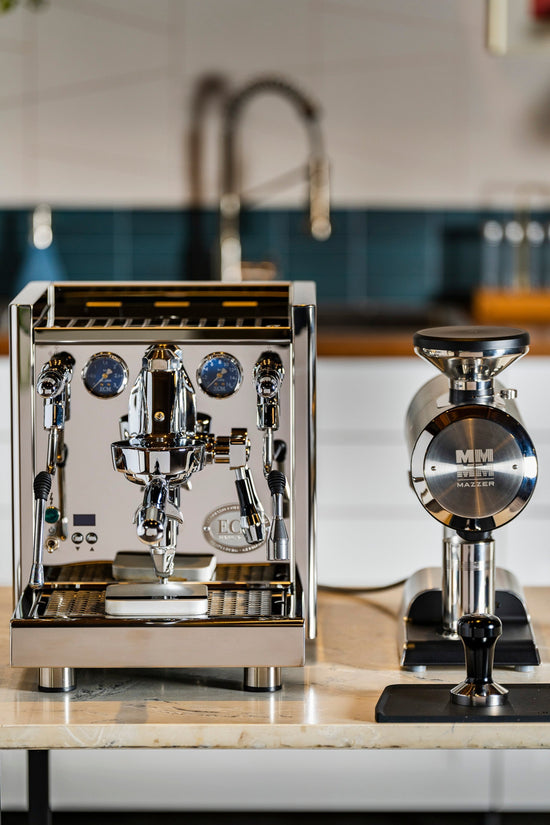 Stainless espresso machine with chrome levers and gauges on a kitchen counter beside a grinder.