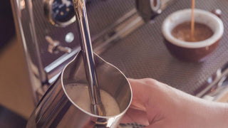 Hand holding a stainless steel frothing pitcher under the steam wand of an espresso machine.