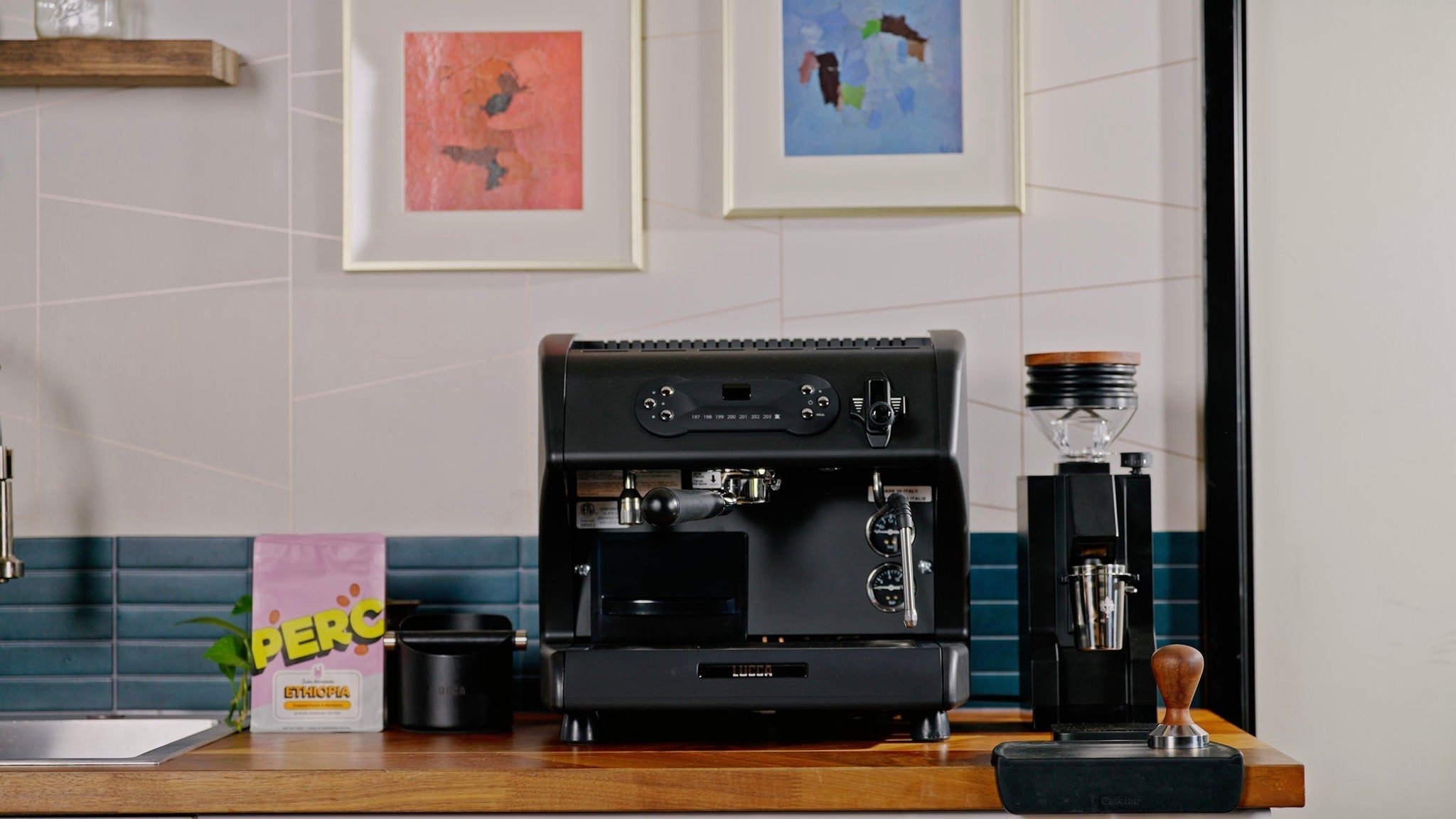 Black Lucca espresso machine on a wooden kitchen counter with abstract framed art on a tiled wall behind and a grinder to the right.