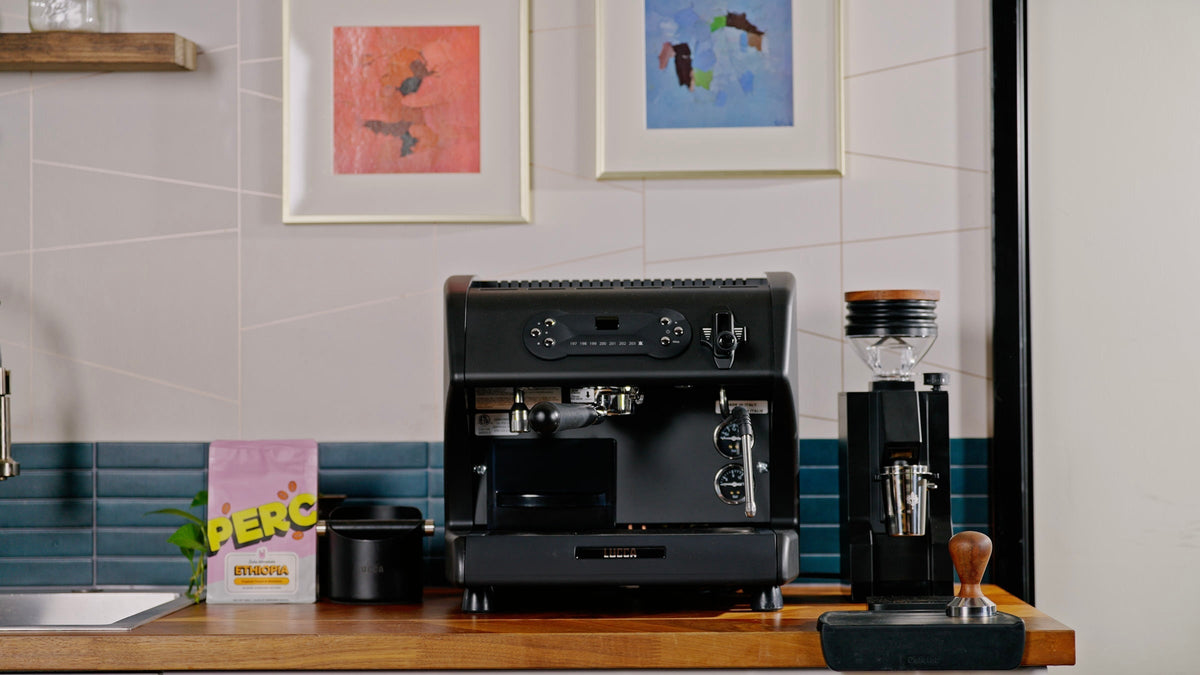 Front view of the LUCCA A53 Pro espresso machine in matte black with an entiire espresso setup lifestyle image by clive coffee