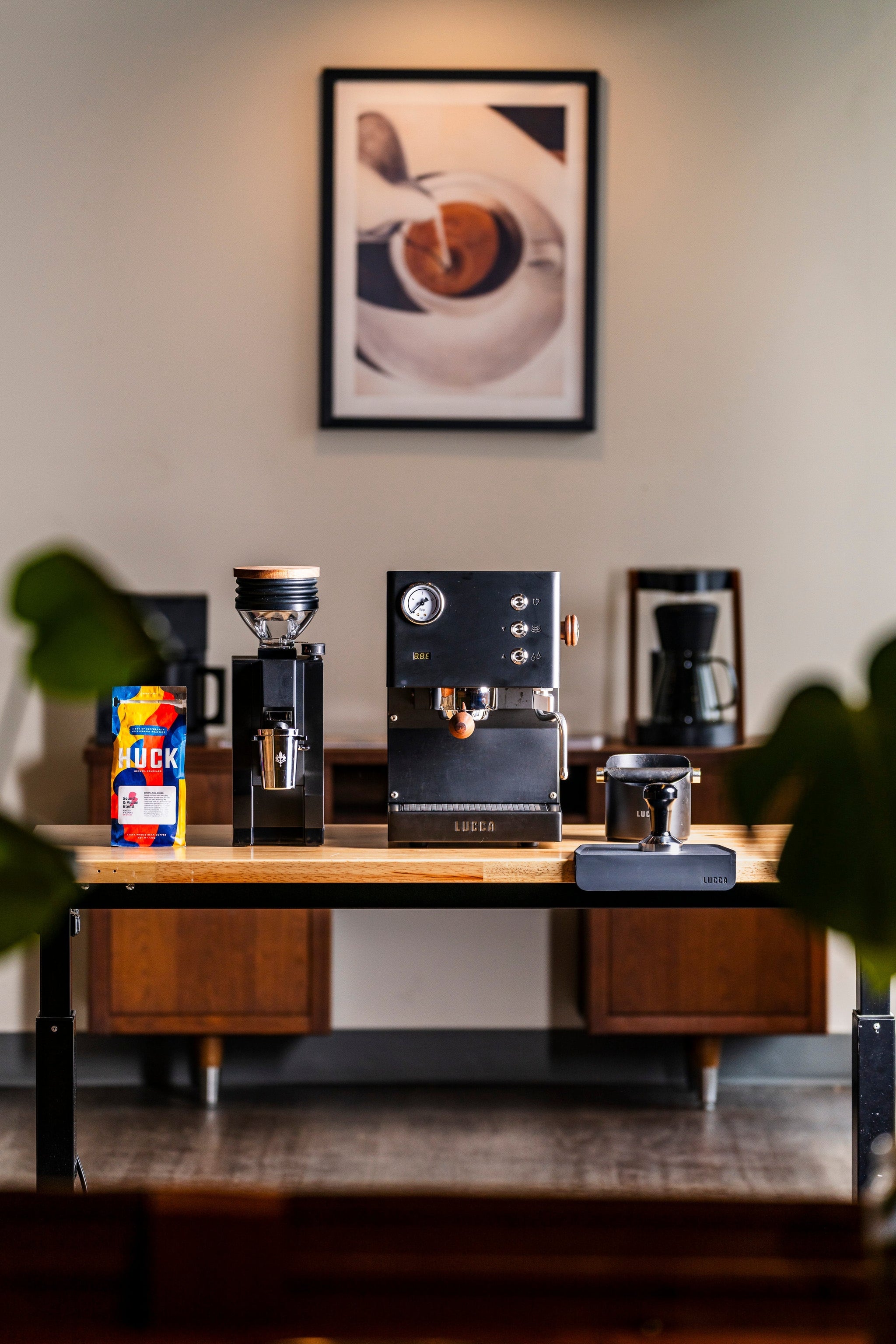 LUCCA espresso machine on a wooden table with a colorful HUCK coffee bag to the left and framed art on the wall behind
