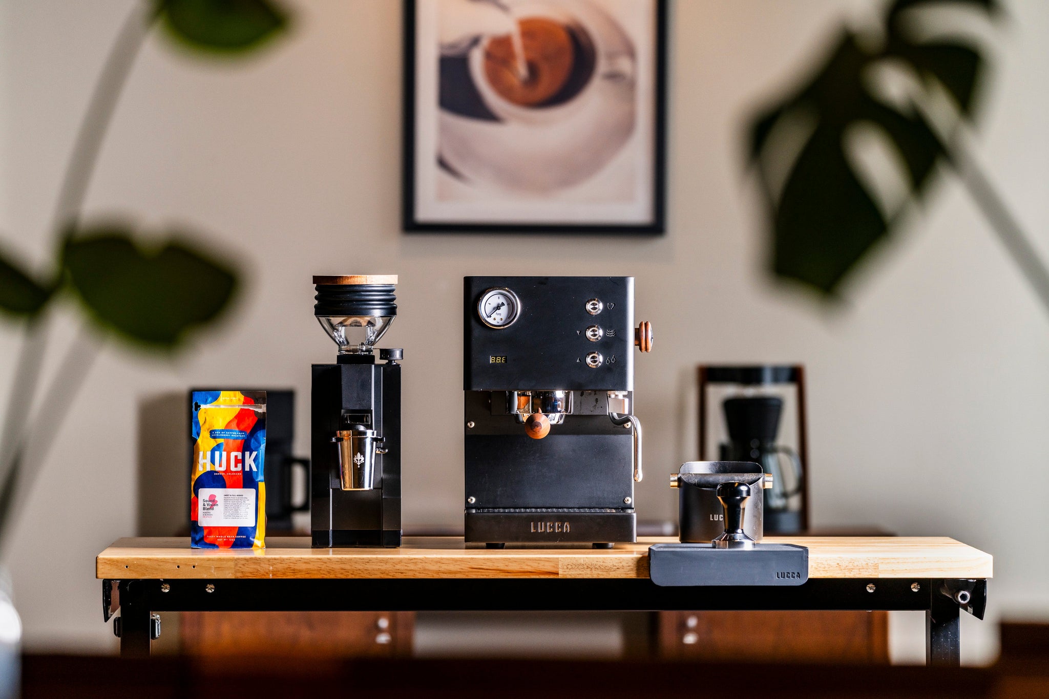Lucca espresso machine on a wooden counter with a coffee bag, a plant, and a framed picture in the background.