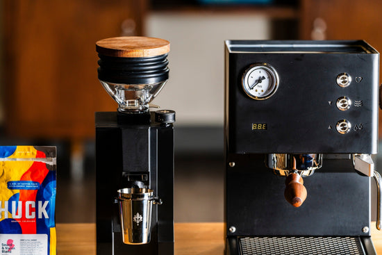 Close up view of the Eureka Zero Mignon Zero 65 Espresso grinder in matte black sitting to the left of a matte black LUCCA Tempo espresso mahcine lifestyle by Clive Coffee