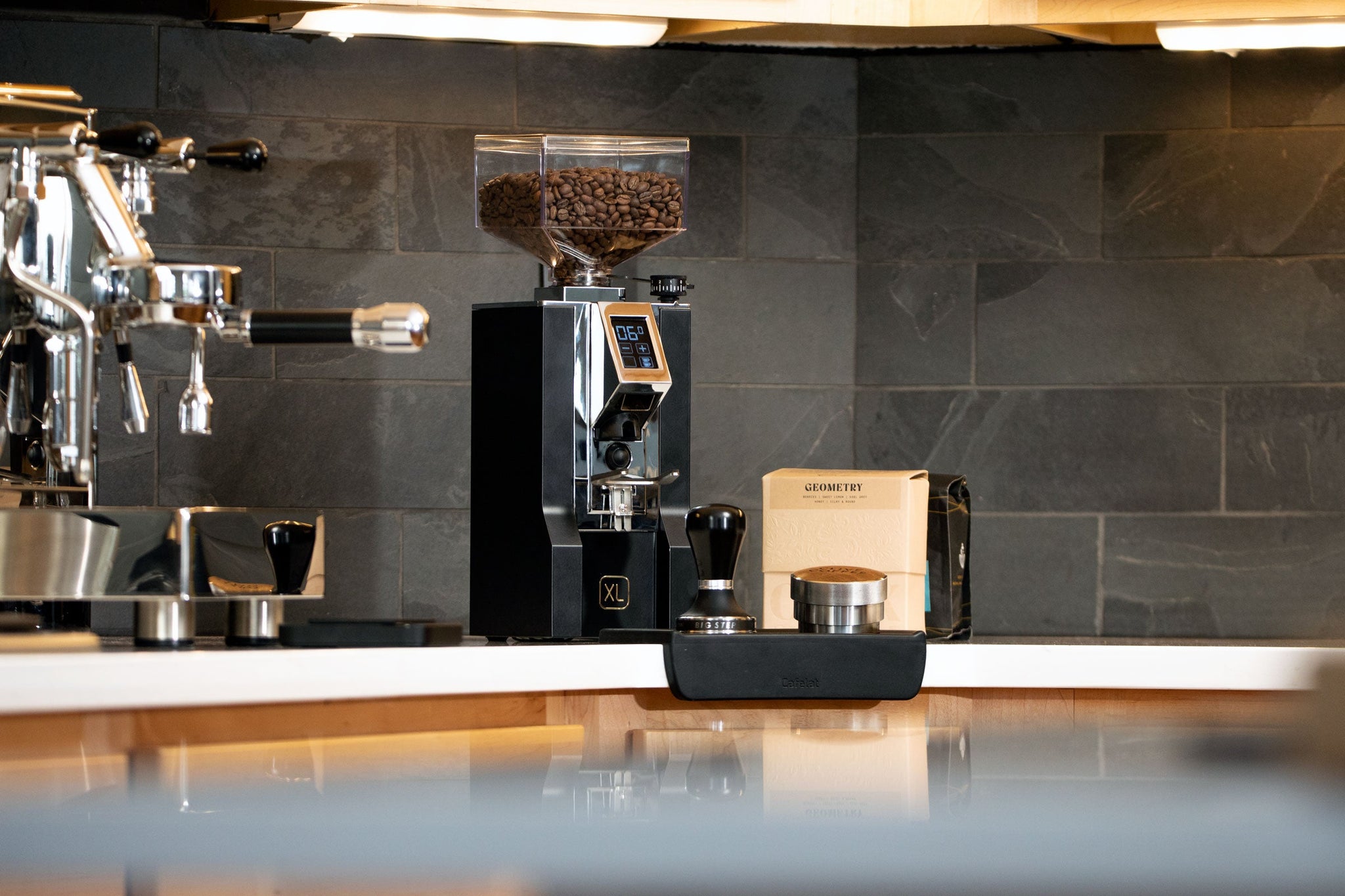 Burr coffee grinder with a clear hopper filled with roasted beans on a kitchen counter beside an espresso machine