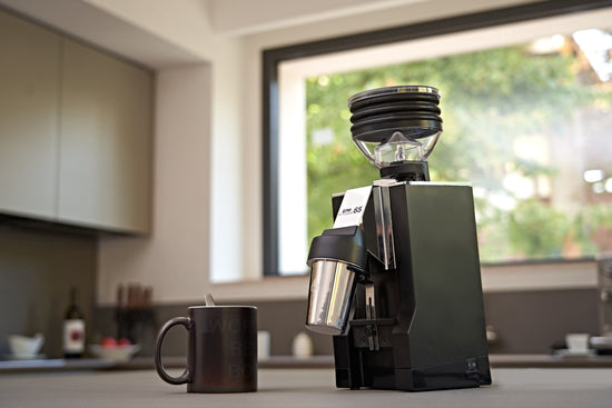 Bright kitchen with a tall black espresso grinder featuring a clear bean hopper beside a window.