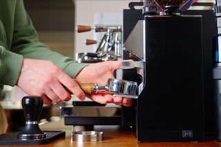 Hands hold a wooden portafilter under a glossy espresso machine, with a tamper on the counter nearby.