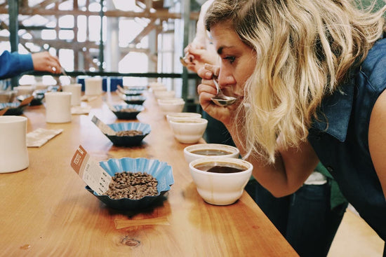 Woman leaning over a long wooden coffee tasting table smelling a cup as bowls of roasted beans and sample cups line the surface.