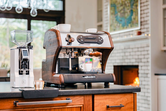 Compact espresso machine with wood side panels and chrome front, two cups on top on a dark kitchen counter.
