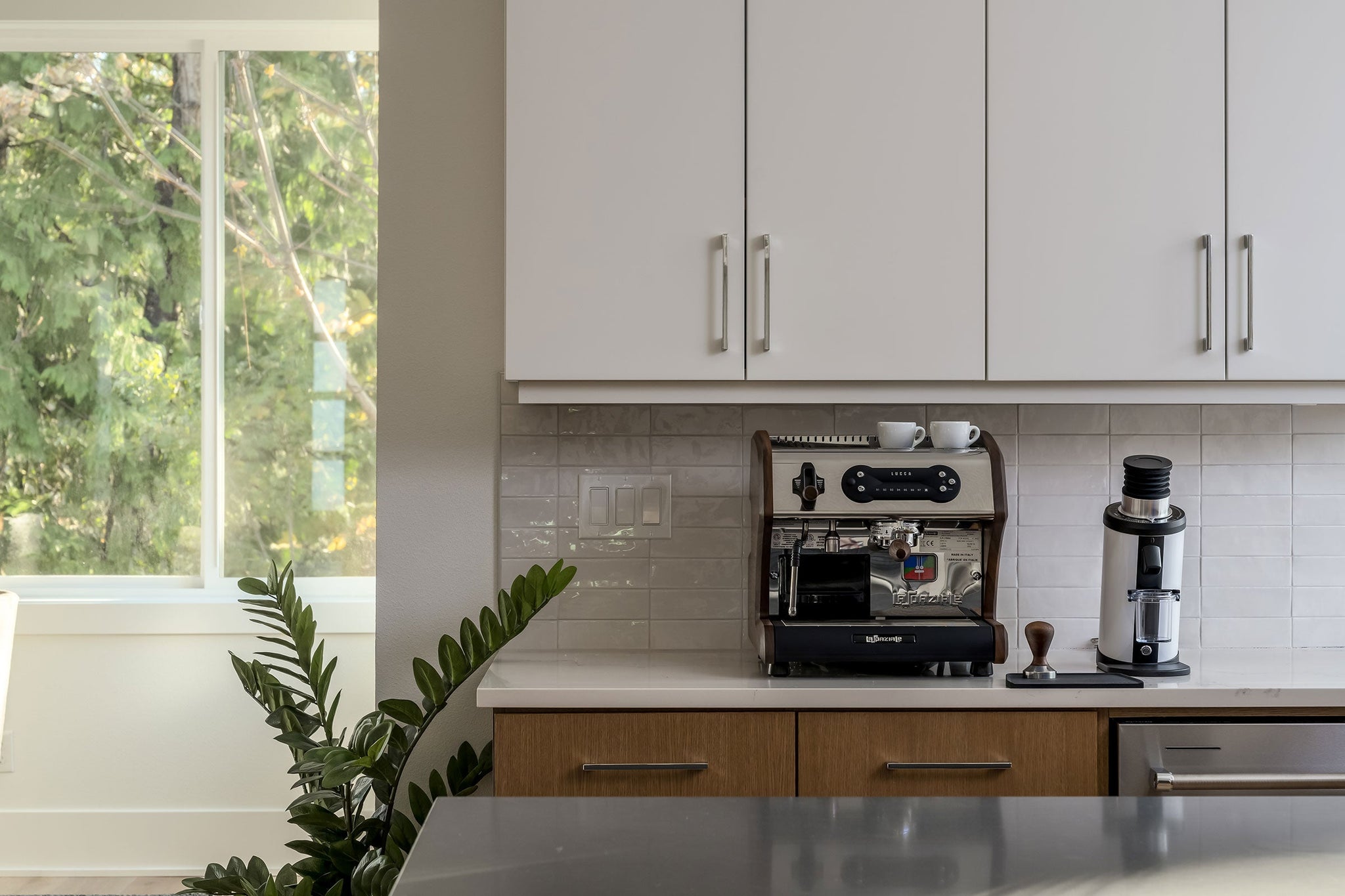 Espresso machine on a white kitchen counter beneath upper cabinets with a leafy window view