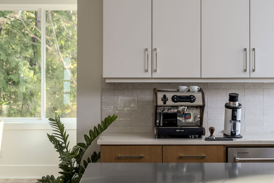 Espresso machine on a white kitchen counter beneath upper cabinets with a leafy window view