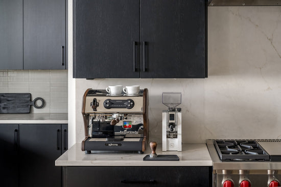 Compact espresso setup under dark kitchen cabinets with two white cups on top