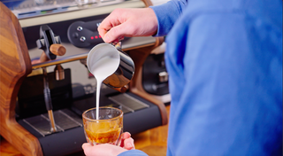 Barista wearing a blue sleeve pours steamed milk from a pitcher into a glass of espresso
