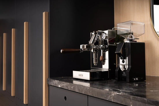 Modern coffee setup featuring LUCCA espresso machine and grinder on a wooden table with framed abstract art above and a bag of HUCK coffee nearby