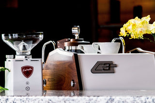 La Marzocco GS3 espresso machine on a counter with a grinder and cups against a dark backdrop.