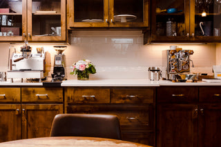 Espresso setup across a wooden kitchen counter with two machines, a grinder, and a flower vase under warm lighting.